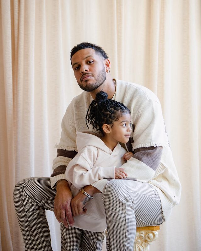 Sarunas J. Jackson in a white t-shirt and black checked white pants with his daughter in same dress.